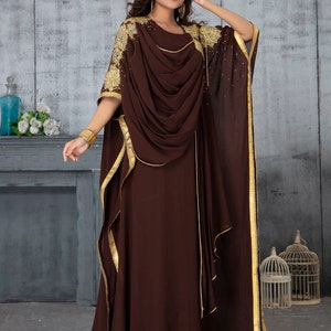 May include: A brown maxi dress with gold trim and embroidery. The dress has a flowing, draped design and a long, flowing skirt.