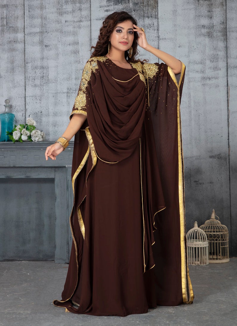 May include: A brown maxi dress with gold trim and embroidery. The dress has a flowing, draped design and a long, flowing skirt.