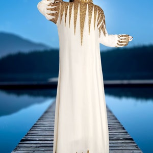May include: A white maxi dress with a gold glitter design. The dress has a high neckline and long sleeves. The dress is decorated with a geometric pattern of gold glitter triangles.