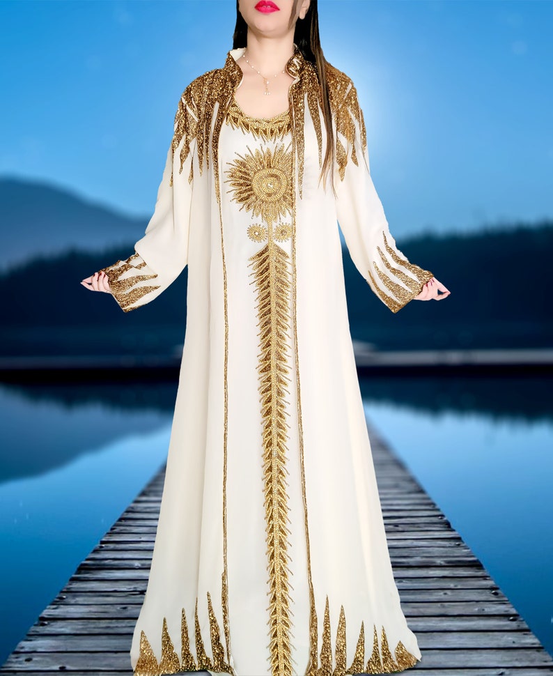 May include: A white maxi dress with gold embroidery. The dress has a long sleeve and a high neckline. The embroidery is intricate and features a floral design.