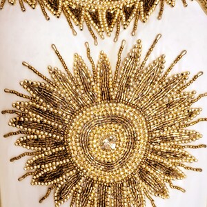 May include: A white garment with gold beading in the shape of a sunburst. The center of the sunburst has a large, clear rhinestone.