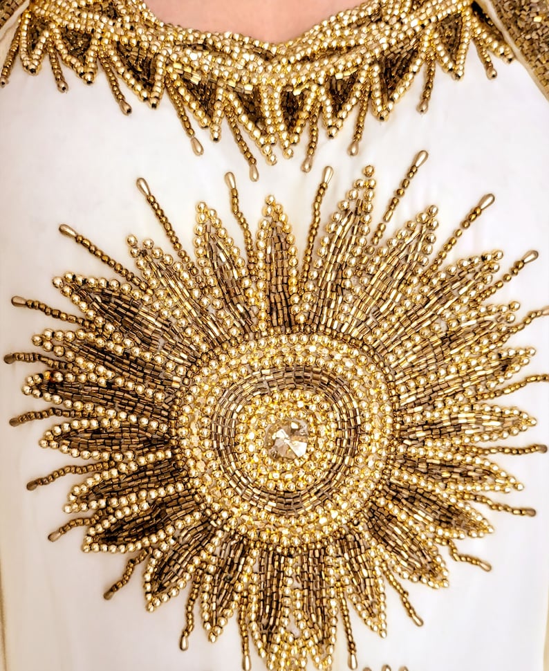 May include: A white garment with gold beading in the shape of a sunburst. The center of the sunburst has a large, clear rhinestone.