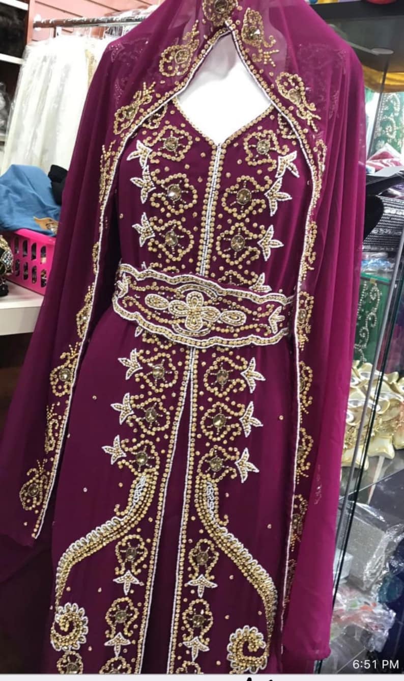 May include: A long, burgundy dress with gold and white beading. The dress has a high neckline and long sleeves. The dress is decorated with intricate floral patterns.