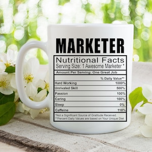 May include: White ceramic mug with a black and white graphic of a nutritional facts label for a "Marketer". The label lists the daily value of hard working, unrivaled skill, passion, caring, sleep, and caffeine.