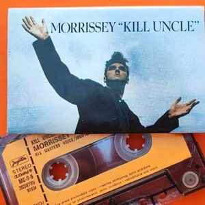 Könnte beinhalten: Eine Kassette mit einem gelben Etikett und einem schwarzen Gehäuse. Das Etikett trägt den Text "KILL UNCLE" und "MORRISSEY" in Schwarz. Die Kassette liegt auf einem größeren, schwarz-weißen Bild von Morrissey, einem Sänger, mit erhobenen Armen.
