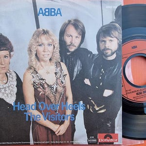 May include: A vinyl record with a black label and an orange centre. The label has the text "ABBA" and "Head Over Heels" printed on it. The record is surrounded by a white paper sleeve with a photo of the band ABBA. The sleeve has the text "Head Over Heels" and "The Visitors" printed on it.