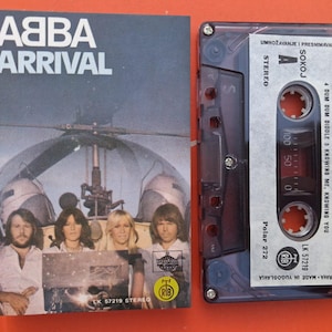 Op de afbeelding: ABBA "Arrival" cassetteband en albumhoes. De hoes toont de band voor een helikopter. De cassetteband is doorschijnend met tracklijsten en stereo-informatie. De albumhoes is blauw met witte tekst.