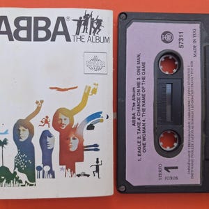 以下が含まれることがあります： ABBAの「The Album」カセットテープとアルバムカバー。カバーには、バンド名とアルバムタイトルが黒で表示され、バンドメンバーのカラフルなイラストが描かれています。カセットは黒色で、紫色のラベルが付いています。