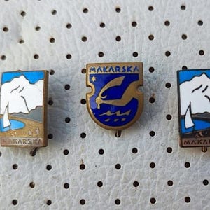 Può includere: Tre spille vintage in smalto con il nome "Makarska" e diversi design. Una spilla presenta una barca a vela di fronte a una montagna, un'altra presenta un delfino in un'onda blu e la terza presenta una barca a vela di fronte a una montagna con una nuvola bianca.