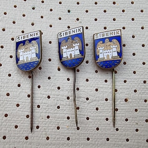 Può includere: Tre spille dorate con smalto blu e dettagli bianchi. Ogni spilla presenta un castello bianco con un'aquila dorata in cima e la parola "SIBENIK" in lettere dorate.