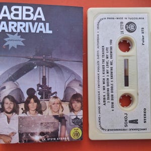 Pode incluir: Fita cassete e capa do álbum "Arrival" do ABBA. A capa apresenta a banda em frente a um helicóptero. A cassete é branco sujo com os títulos das músicas. Um item de colecionador de música retrô.