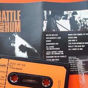 Op de afbeelding: Een oranje cassettebandje en albumhoes van U2's "Rattle and Hum". De albumhoes is zwart-wit met oranje tekst. Het cassettebandje is oranje met zwarte tekst.