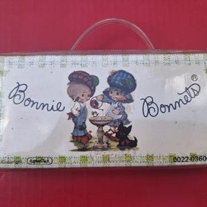 May include: A rectangular product package labelled Bonnie Bonnets with a clear plastic handle. The label features a graphic of two children in plaid hats at a birdbath with a small black dog. The border has a light green and white checkered pattern.