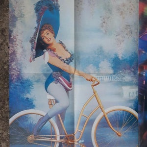May include: A vintage poster featuring Marilyn Monroe in a blue and white dress riding a gold bicycle with white tyres. The poster has a blue and white background with a text overlay that reads "marilyn monroe".