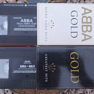 Könnte beinhalten: Zwei ABBA VHS-Kassetten und zwei Kassettenhüllen. Die Kassetten sind schwarz mit weißem Text, und die Hüllen sind weiß und schwarz mit goldfarbenen Buchstaben. Die Kassetten und Hüllen zeigen das ABBA Gold Album.