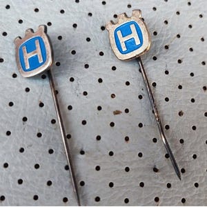 Peut inclure: Deux épingles de revers Husqvarna vintage. Chaque épingle présente un emblème argenté avec un fond bleu et un "H" blanc au centre. Les épingles ont une épingle droite argentée attachée à l'emblème.