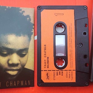 Op de afbeelding: Een cassettebandje met een oranje label met de tekst "TRACY CHAPMAN" en "WEA/JOGOTON". De cassettebandje zit in een zwarte plastic cassette.