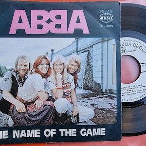 Könnte beinhalten: Eine Vintage-45-RPM-Single der schwedischen Popgruppe ABBA. Die Schallplatte enthält den Song "The Name of the Game". Das Cover zeigt die vier Mitglieder der Gruppe bei einem Fotoshooting vor einem Wasserfall.
