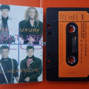 Op de afbeelding: Een cassettebandje met een oranje label. Het label heeft de tekst "From Luxury to Heartache" en "Culture Club" in zwarte tekst. De cassetteband is zwart met een zilveren metalen strip.