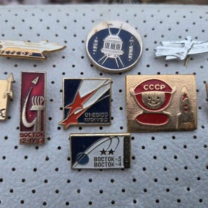 Puede incluir: Una colección de insignias y pines vintage con temática espacial. Los pines presentan varios diseños, incluyendo cohetes, satélites y el logo CCCP. Algunos pines tienen texto como "BOCTOK" y "1959". La paleta de colores incluye dorado, rojo, azul y blanco.