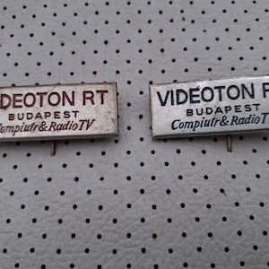 Könnte beinhalten: Zwei rechteckige Metallabzeichen mit der Aufschrift "VIDEOTON RT BUDAPEST Compuiutr & Radio TV". Eines hat roten Text, das andere blauen Text. Jedes Abzeichen hat eine kleine Nadel auf der Rückseite.