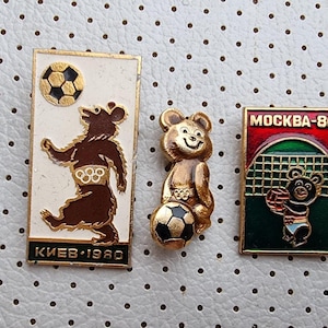 Op de afbeelding: Drie vintage emaille-speldjes met een beer met een voetbal, een beer met een voetbal en de tekst "KIEV 1980", en een beer met een voetbal en de tekst "MOCKBA-80".