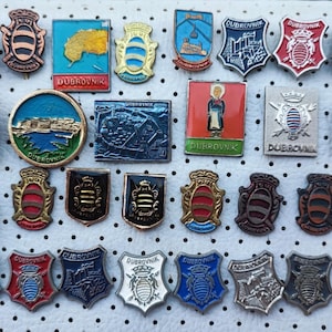 Può includere: Una collezione di 30 spille smaltate vintage che raffigurano lo stemma di Dubrovnik, Croazia. Le spille sono di vari colori e design, tra cui rosso, blu, verde e oro. Alcune spille mostrano il nome della città, "Dubrovnik", mentre altre mostrano lo stemma della città, che include uno scudo con un leone e una corona.