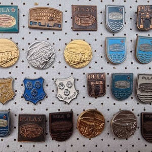 Può includere: Una collezione di spille smaltate vintage con vari disegni, tra cui un anfiteatro romano, una porta della città e uno scudo con una testa di leone. Le spille sono disposte su uno sfondo bianco con un motivo a griglia. Le spille sono di vari colori, tra cui blu, rosso, oro e argento.