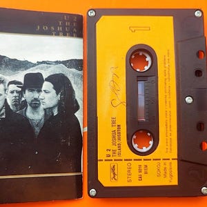 Op de afbeelding: Een vintage cassettebandje van U2's album "The Joshua Tree" met een geel label en zwarte behuizing. De albumhoes toont een zwart-wit foto van de band. De cassettehoes is open en toont de band en album artwork.