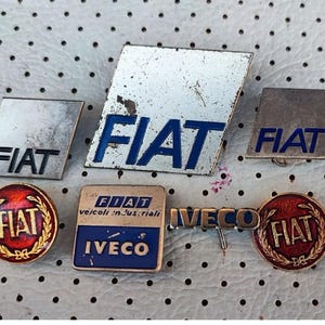 以下が含まれることがあります： ヴィンテージのフィアット車のエンブレムとバッジのコレクション。黒と青のフィアットのロゴが入った様々な形とデザインが含まれています。一部のバッジは、赤と金の配色で丸くなっています。