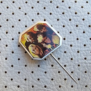 Könnte beinhalten: Eine silberne Anstecknadel mit einem bunten Bild der Band ABBA. Die Nadel befindet sich auf einem weißen Hintergrund mit kleinen Löchern.