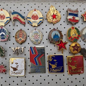 Pode incluir: Uma coleção de alfinetes e distintivos vintage, apresentando vários designs com estrelas vermelhas, bandeiras e emblemas. Alguns alfinetes têm texto, e a paleta de cores inclui vermelho, dourado, azul e branco.