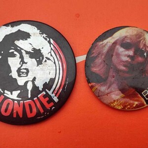 Op de afbeelding: Twee ronde, zwart-witte, pinback-knoppen met een afbeelding van een vrouw met blond haar en het woord "BLONDIE" in rode letters.