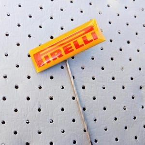 Könnte beinhalten: Ein gelb-roter Pirelli-Ansteckpin mit dem Pirelli-Logo. Der rechteckige Pin hat eine silberne Nadel. Er befindet sich auf einem hellblauen Hintergrund mit kleinen, gleichmäßig verteilten Löchern, ähnlich einem Autositz.
