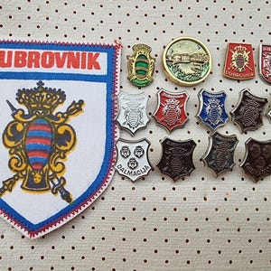 Può includere: Una collezione di spille smaltate vintage con vari disegni, tra cui uno scudo con la parola "DUBROVNIK" e un'ape, uno scudo con la parola "DALMACIJA" e tre leoni, e uno scudo con la parola "DUBROVNIK" e un'ape. Le spille sono disposte in fila su una superficie bianca con un motivo a punti.