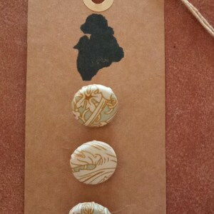 May include: A brown paper tag featuring a black silhouette and three fabric-covered buttons. The buttons display a cream and light blue floral pattern. A natural twine string is attached.
