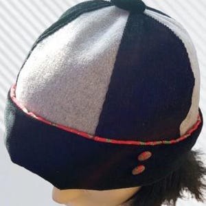 May include: A colour-blocked hat with a black brim, a grey and white panel, and a black top. The brim is trimmed with a red and green patterned ribbon. Two small, round, brown buttons are on the side of the hat.