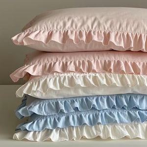 May include: A stack of four ruffled pillowcases in various shades of pink and blue. The top pillowcase is a light pink, the second is a darker pink, the third is white, and the bottom is a light blue. All the pillowcases are trimmed with a ruffled edge.