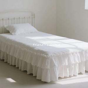 May include: A white bed with a frilled valance and a matching pillow. The bed frame is a light cream colour with a decorative headboard. The bed is in a well-lit room.