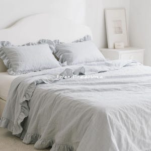May include: A bed with a white headboard and light blue bedding. The bedding includes a ruffled duvet cover and matching pillowcases. The room is bright and airy, with a framed picture on a white nightstand.