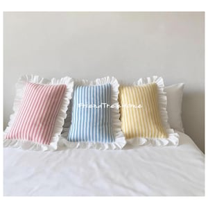 Puede incluir: Tres cojines decorativos a rayas en rosa, azul y amarillo. Cada cojín tiene un borde blanco con volantes. Los cojines están colocados sobre una cama blanca.