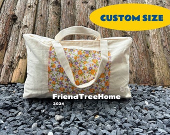 May include: A white canvas tote bag with a floral print insert. The bag has two handles and the text "FriendTreeHome 2024" is printed on the bottom. The bag is sitting on a bed of small gray rocks. A yellow oval with the text "CUSTOM SIZE" is in the top right corner of the image.