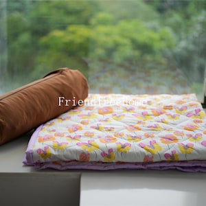 May include: A white and purple floral patterned quilt with a brown cylindrical pillow on top. The quilt is folded in half and the pillow is lying on top of the folded quilt. The quilt and pillow are sitting on a white surface.