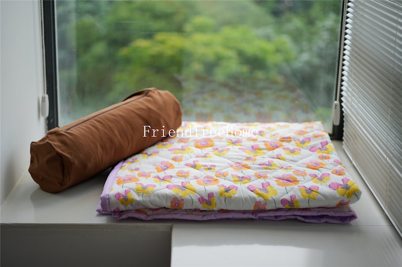 May include: A white and purple floral patterned quilt with a brown cylindrical pillow on top. The quilt is folded in half and the pillow is lying on top of the folded quilt. The quilt and pillow are sitting on a white surface.
