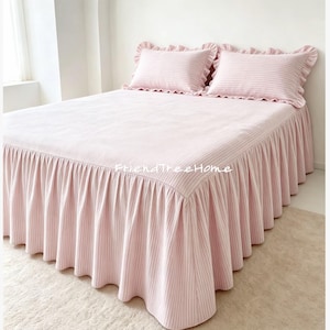 May include: A light pink bedspread with a white vertical stripe pattern, featuring a ruffled skirt and two matching ruffled pillow shams. The text FriendTreeHome is printed in white on the center of the bedspread surface.