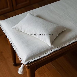 May include: A white linen bench cushion with a white fringe border and a white pillow on top. The cushion is on a wooden bench.