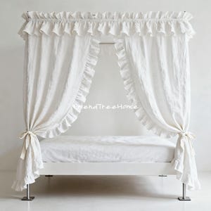 Puede incluir: Una cama con dosel blanca con adornos y cortinas con volantes. El marco de la cama es blanco y las cortinas están atadas con lazos. La cama está cubierta con ropa de cama blanca.