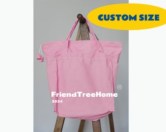 May include: A pink canvas tote bag with a drawstring closure and the text "FriendTreeHome 2024" printed on the front. The bag is sitting on a wooden stand. The text "CUSTOM SIZE" is printed in a yellow box above the bag.