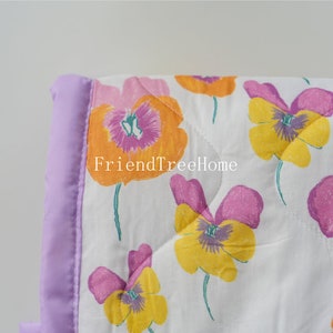 May include: A white quilt with a purple satin border and a floral pattern of yellow, pink, and orange pansies. The quilt has a quilted diamond pattern. The text "FriendTreeHome" is visible in the image.