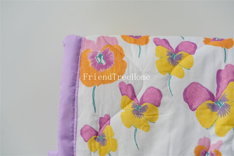 May include: A white quilt with a purple satin border and a floral pattern of yellow, pink, and orange pansies. The quilt has a quilted diamond pattern. The text "FriendTreeHome" is visible in the image.
